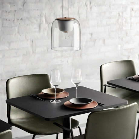 "A modern pendant light with a clear glass shade and wooden top, hanging above a dining table with place settings and wine glasses."