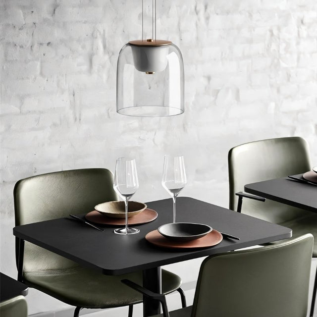 "A modern pendant light with a clear glass shade and wooden top, hanging above a dining table with place settings and wine glasses."