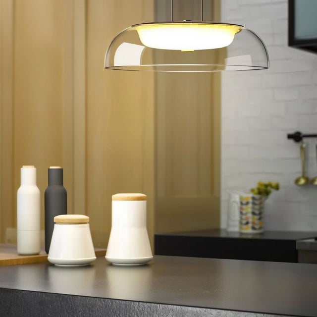 "A modern pendant light with a clear glass dome shade, hanging above a kitchen counter with ceramic containers and bottles."