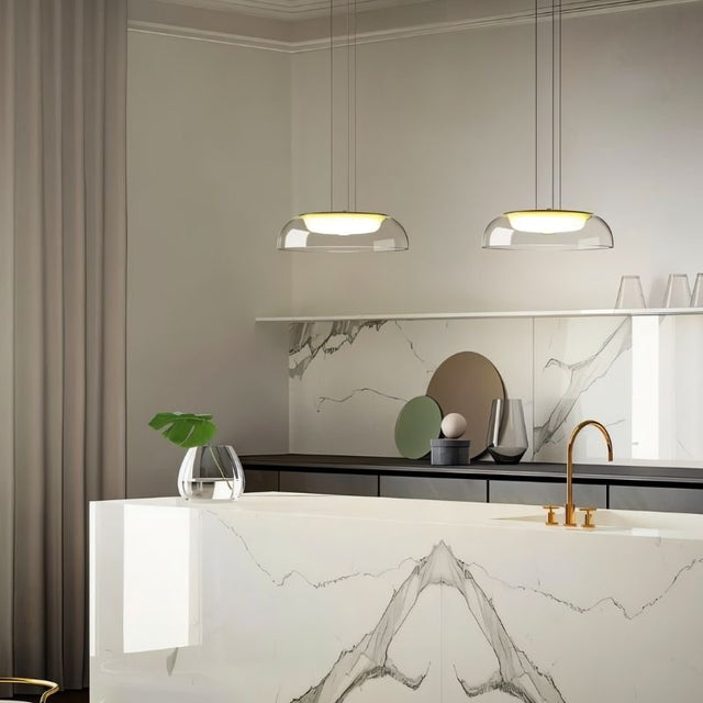 "Two modern pendant lights with clear glass dome shades, hanging above a kitchen island with a marble countertop and a glass vase."