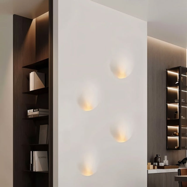 "Four white gypsum Onexa trimless wall lights with dome-like profiles, casting ambient light in a modern interior with built-in shelving."