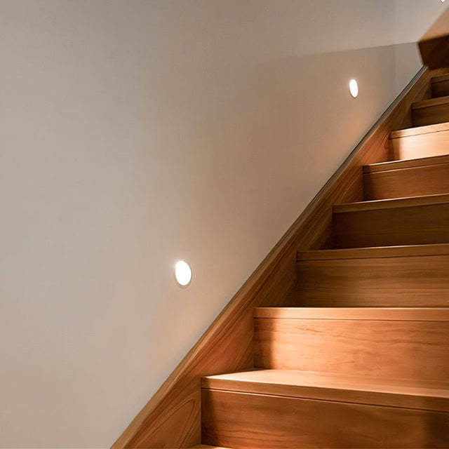 "Two white gypsum Onexa trimless wall lights with rounded cutouts, casting ambient light on a wall beside wooden stairs."