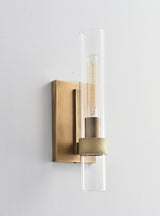 "A modern brass wall lamp with a cylindrical glass shade, blending vintage and contemporary styles with a bronze finish and black accents."