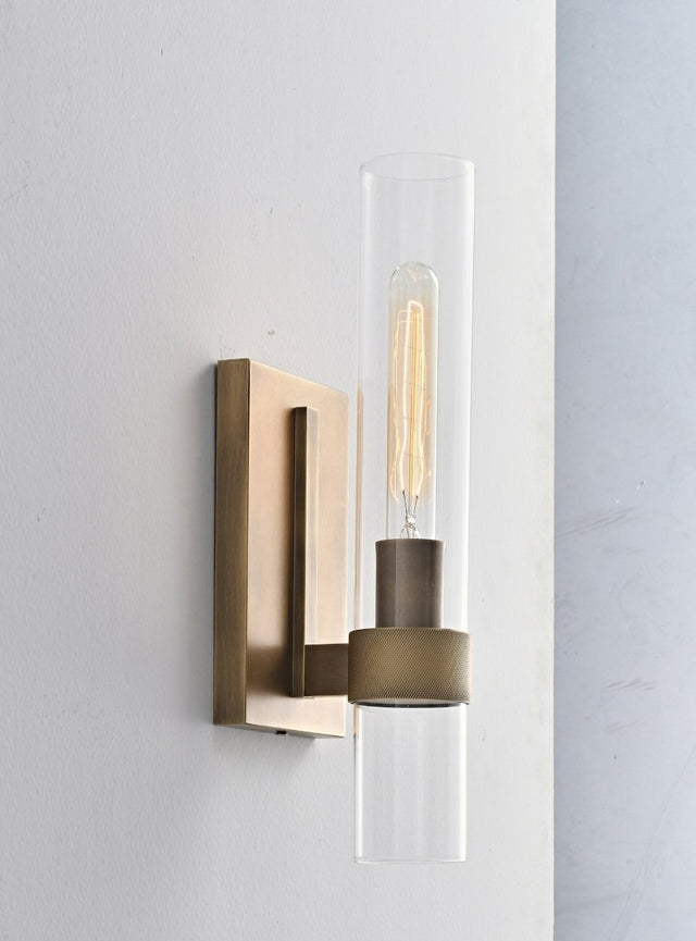 "A modern brass wall lamp with a cylindrical glass shade, blending vintage and contemporary styles with a bronze finish and black accents."