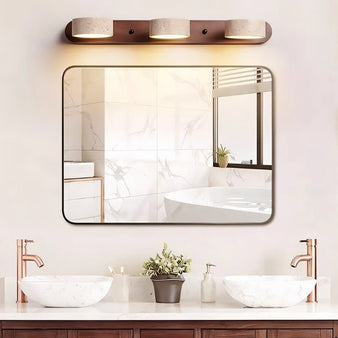 "A minimalist three-light travertine vanity light on a dark wood base, illuminating a modern bathroom mirror and sink."