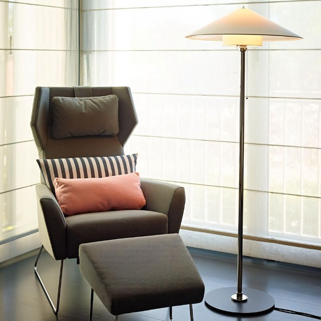 "The Oriel floor lamp with a sleek metallic frame and wide conical fabric shade, placed next to a modern armchair, made from aluminum, iron, and fabric, providing soft, diffused lighting in a stylish living room setting."