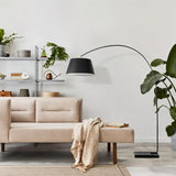 "A modern arc floor lamp with a black metal frame and a black fabric shade stands next to a beige sofa in a living room with plants and shelving in the background."