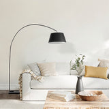 "A modern arc floor lamp with a black metal frame and black fabric shade illuminates a white sofa in a living room with a coffee table and decorative items."