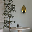 "Lit copper wall lamp with a leaf-inspired design mounted on a wall in a room with a plant and table."