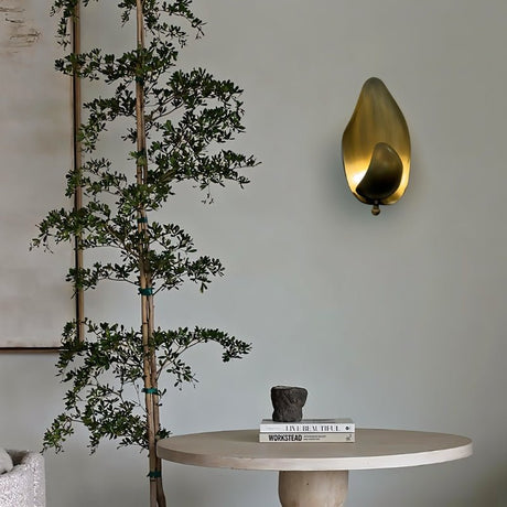 "Lit copper wall lamp with a leaf-inspired design mounted on a wall in a room with a plant and table."
