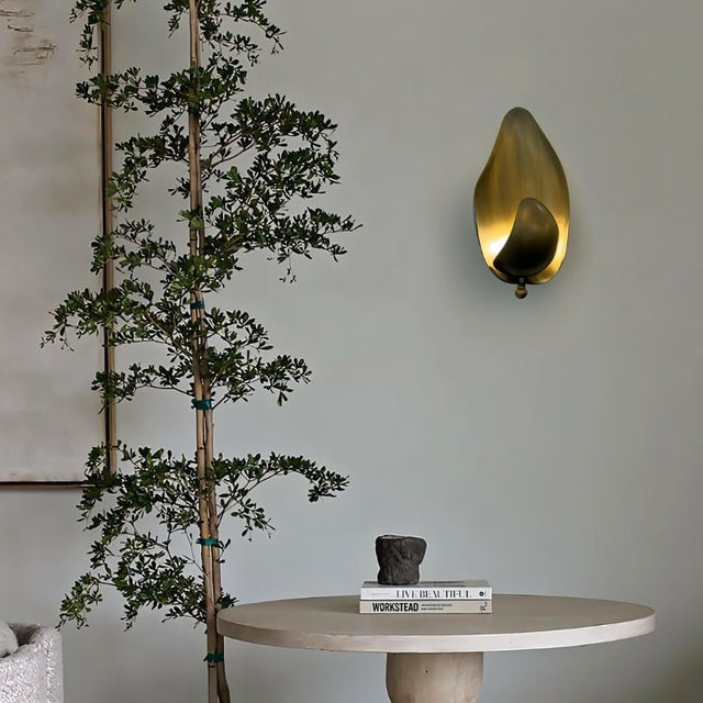 "Lit copper wall lamp with a leaf-inspired design mounted on a wall in a room with a plant and table."