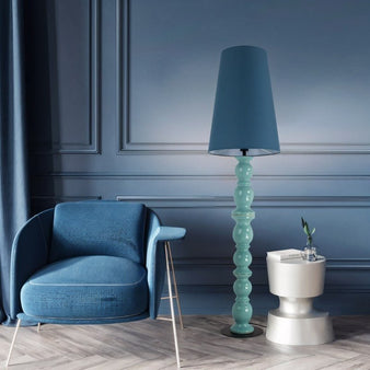 "A floor lamp with a blue sculptural ceramic base and a tall, tapered shade in a modern living room with a matching blue chair."