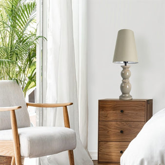 "A taupe table lamp with a ceramic base and a tapered cloth shade on a wooden nightstand in a modern bedroom with a large window."