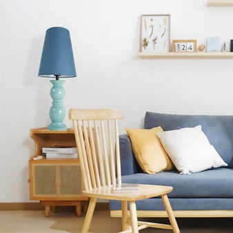 "A teal table lamp with a ceramic base and a tapered cloth shade on a nightstand in a modern bedroom with a blue sofa."