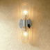 Gold wall sconce with two textured glass cylindrical shades mounted on white bedroom wall above potted plant.
