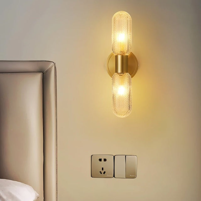"A modern brass wall lamp with textured glass shades above a switch plate in a bedroom."