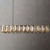 Linear chandelier with brass finish and stacked crystal prisms, suspended by chains against a neutral gray wall.