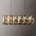Brass linear chandelier with geometric crystal panels suspended from chains against a neutral gray wall.