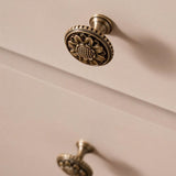 "Antique brass floral knob with detailed floral motif and textured border on a light cabinet drawer."