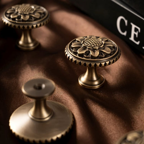 "Set of antique brass floral knobs with detailed floral motif and textured border on a brown fabric."