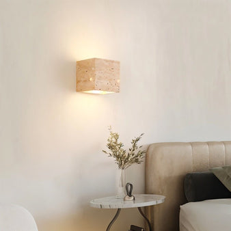 "A modern, cube-shaped wall lamp made of yellow travertine, mounted on a white wall in a bedroom, emitting a soft, ambient glow."