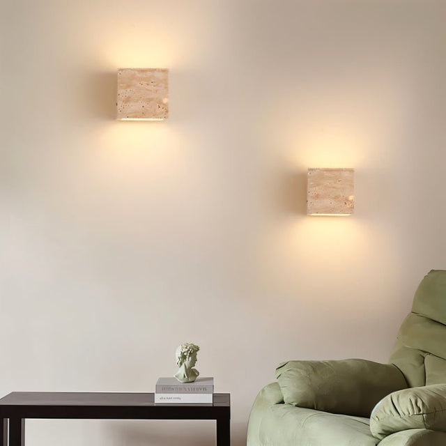 "A pair of modern, cube-shaped wall lamps made of yellow travertine, mounted symmetrically on a neutral wall, emitting a soft, ambient glow in a living room setting."