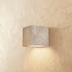 Two cream travertine wall sconces with warm lighting on white wall beside taupe curtains in modern interior space.