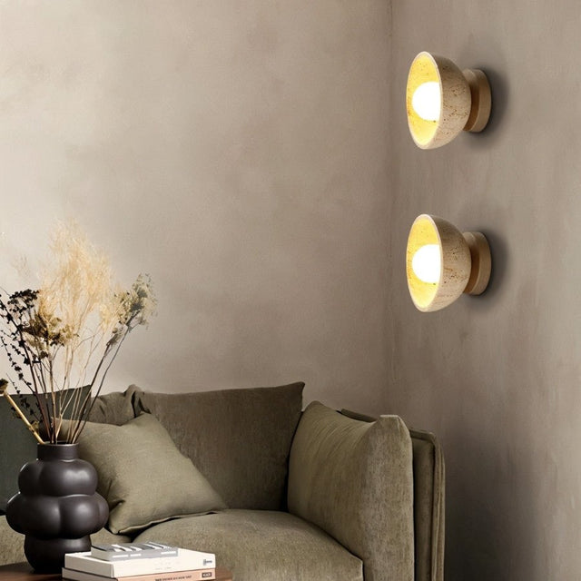 "Pair of travertine wall lamps with circular wooden backplates, illuminating a living room with a comfortable sofa."