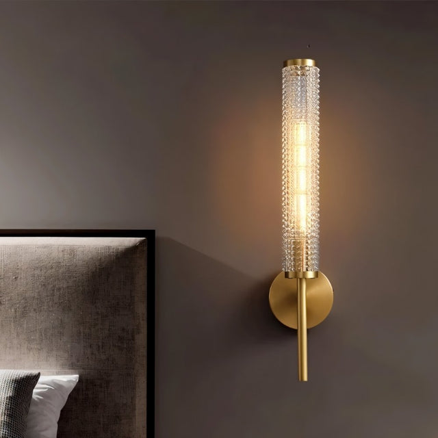 "A modern brass wall lamp with a textured cylindrical glass shade in a bedroom."