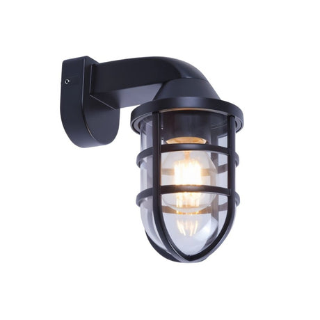 "A brass-colored, industrial-style wall lamp with a robust cage structure and clear PC diffuser, mounted on a stone wall, illuminating a garden path at night."