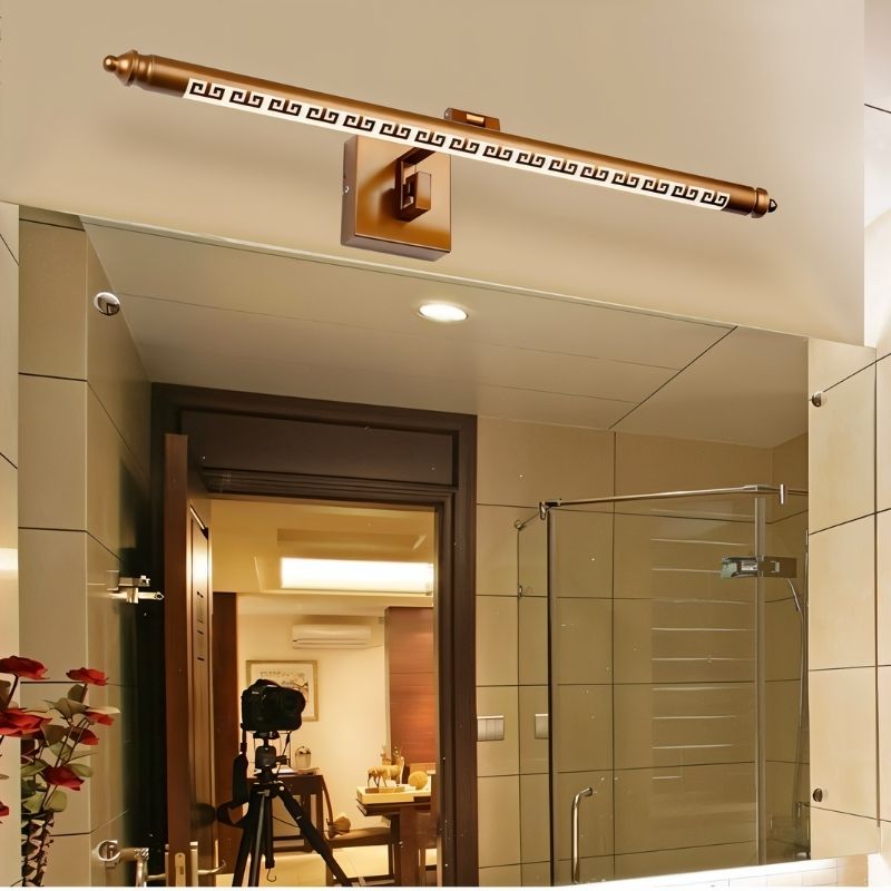 "A gold wall lamp with a Greek key pattern mounted above a bathroom mirror, reflecting an open doorway and a tripod camera."