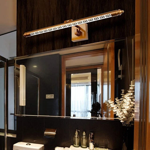 "A gold wall lamp with a Greek key pattern mounted above a bathroom vanity mirror, illuminating the sink and toiletries."