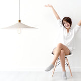 "Raida pendant light with a white conical design, illuminating a modern lifestyle space with a cheerful woman sitting in a minimalist chair, creating a bright and welcoming atmosphere."