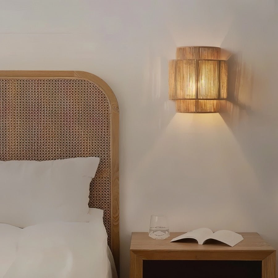 "A bamboo and rattan wall lamp with hemp rope detailing serving as a sconce in a rustic bedroom, casting a warm, patterned light."