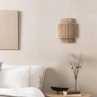 "A layered cylindrical wall lamp made of bamboo, rattan, and hemp rope, diffusing a warm light in a rustic bedroom."