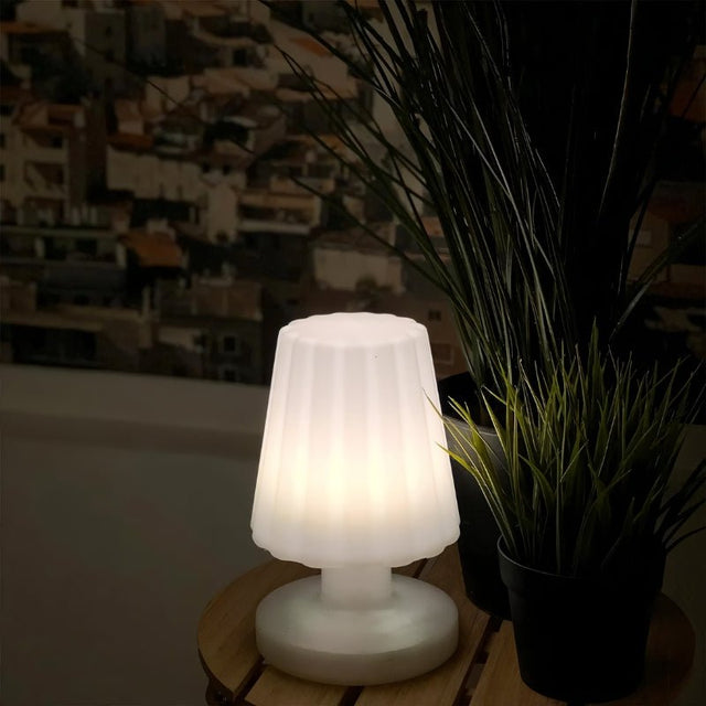 "A compact, cordless table lamp with a soft, fluted shade, casting a warm, inviting glow in a dimly lit setting, with plants in the background."