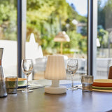 "A modern, cordless table lamp with a soft, fluted shade, illuminating an indoor dining table with place settings and glassware, with a bright, natural light background."
