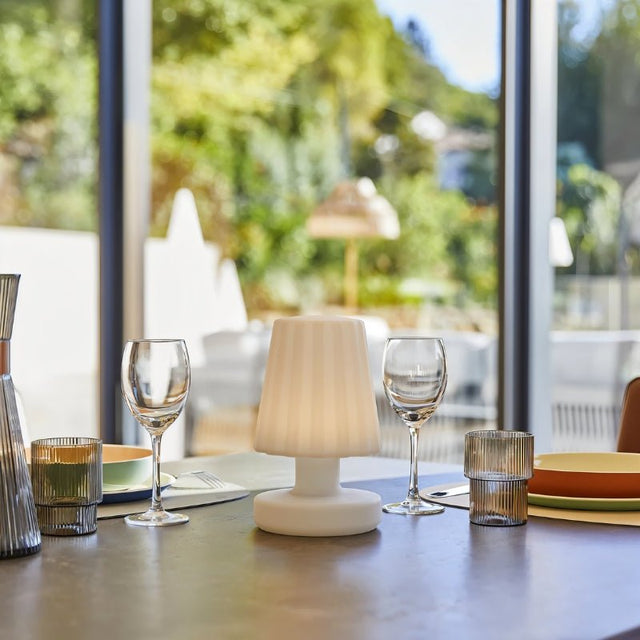 "A modern, cordless table lamp with a soft, fluted shade, illuminating an indoor dining table with place settings and glassware, with a bright, natural light background."