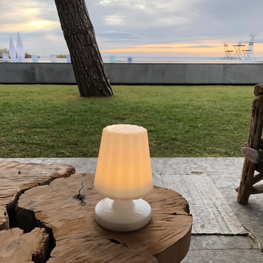 "A compact, cordless table lamp with a fluted shade, casting a warm glow on a rustic outdoor table with a scenic view of a lake and sunset."