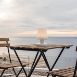 "A compact, cordless table lamp with a soft, fluted shade, illuminating an outdoor balcony table with a scenic ocean view."