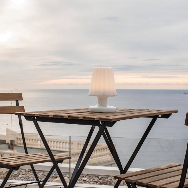 "A compact, cordless table lamp with a soft, fluted shade, illuminating an outdoor balcony table with a scenic ocean view."