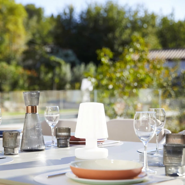 "A compact, cordless table lamp with a fluted shade, providing a warm glow on an outdoor dining table with place settings, with a blurred green background."
