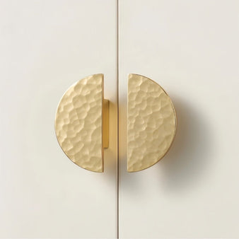 "Pair of hammered brass half-circle knobs with a textured, artisanal finish, mounted on a modern white cabinet."