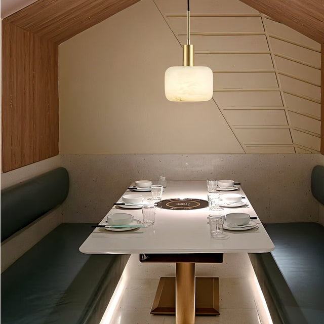 "A modern pendant light with a rounded alabaster shade and brass accents hangs above a dining table set for a meal, providing a soft, warm glow."