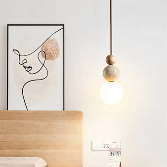 "Sculptural pendant light with stacked travertine, wood, and metal elements, featuring a frosted glass diffuser, hanging in a bedroom."