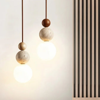 "Two sculptural pendant lights with stacked travertine, wood, and metal elements, featuring frosted glass diffusers."