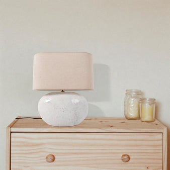 "A modern ceramic table lamp with a beige linen shade, placed on a wooden dresser in a minimalist bedroom, providing a warm and inviting ambiance."