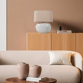 "A modern ceramic table lamp with a linen shade, illuminating a transitional living room with warm beige walls, a neutral sofa, and decorative vases."