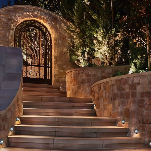 "Sleek, round step lights with clear acrylic diffusers, illuminating a stone stairway leading to an ornate gate and lush greenery."