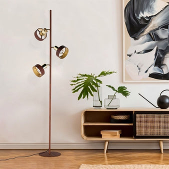 "A modern floor lamp with three adjustable shades in a wood-like and brass finish, illuminating a stylish living room with a contemporary cabinet, artwork, and plants."